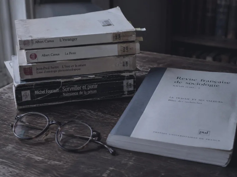 A stack of well worn French paperbacks, Camus, Sartre and Foucault, beside an issue of the Revue française de sociologie and a pair of reading glasses.